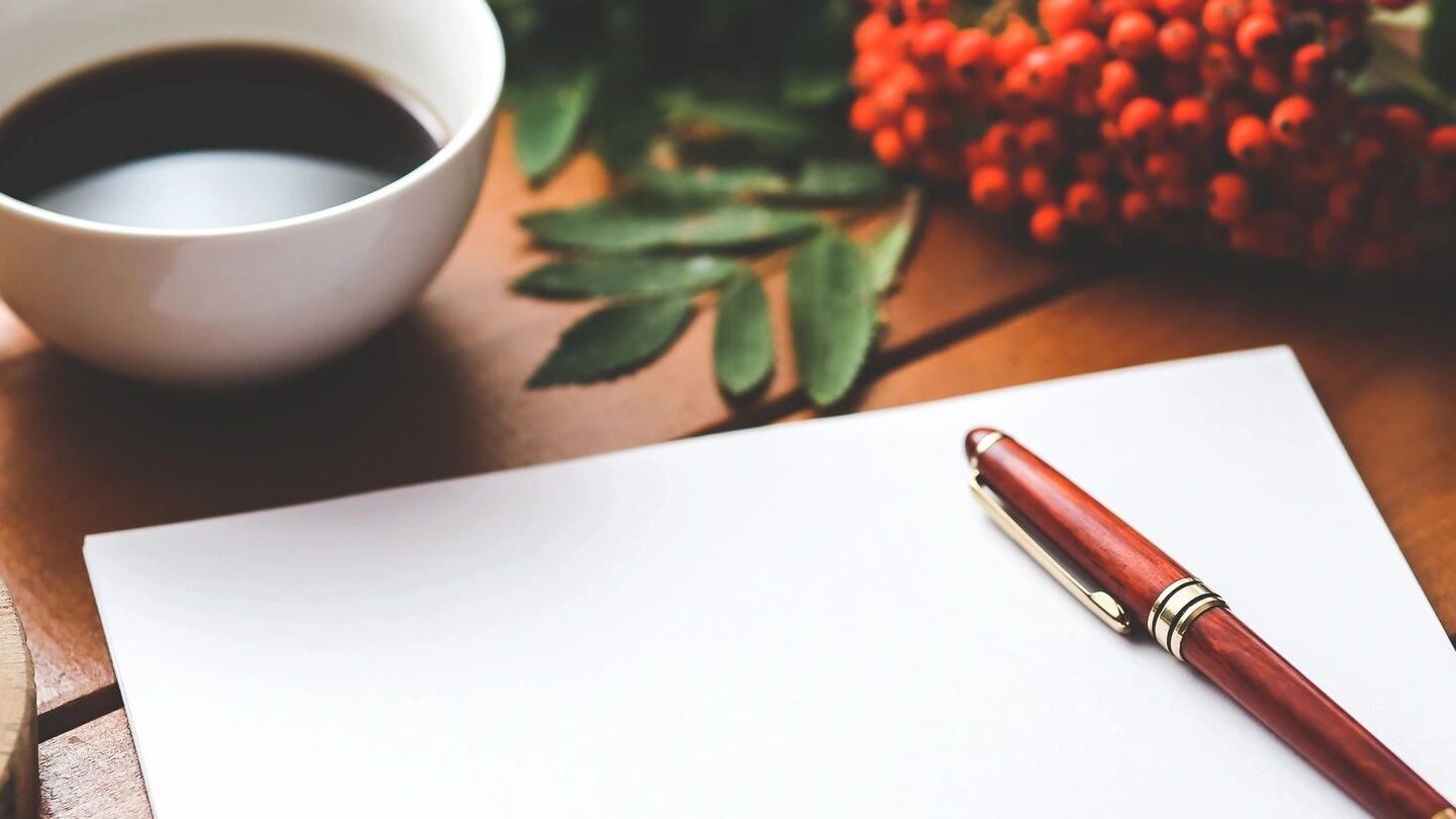 A bronze pen sits on a blank sheet of paper, ready for a story to be told. the page sits atop a wooden surface, a white mug of coffee above it and flowers to the right.