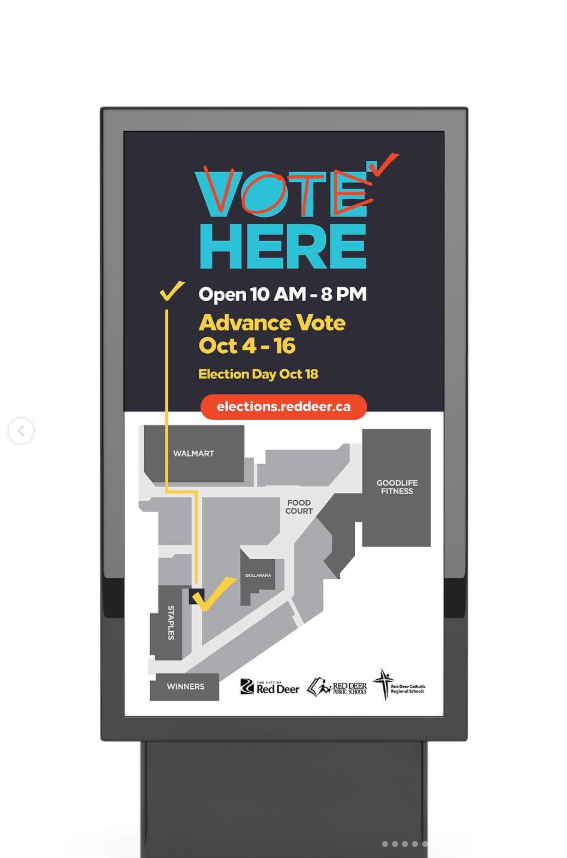 A freestanding digital wayfinding sign for a voting station inside a shopping mall during the City of Red Deer 2021 Election. The screen reads “Vote Here” with hours listed as “Open 10 AM – 8 PM,” along with “Advance Vote Oct 4–16” and “Election Day Oct 18.” Below the text is a simplified mall map showing nearby retailers such as Walmart, Winners, Staples, and GoodLife Fitness, with a highlighted path guiding voters to the polling station. City and school board logos appear along the bottom of the screen.