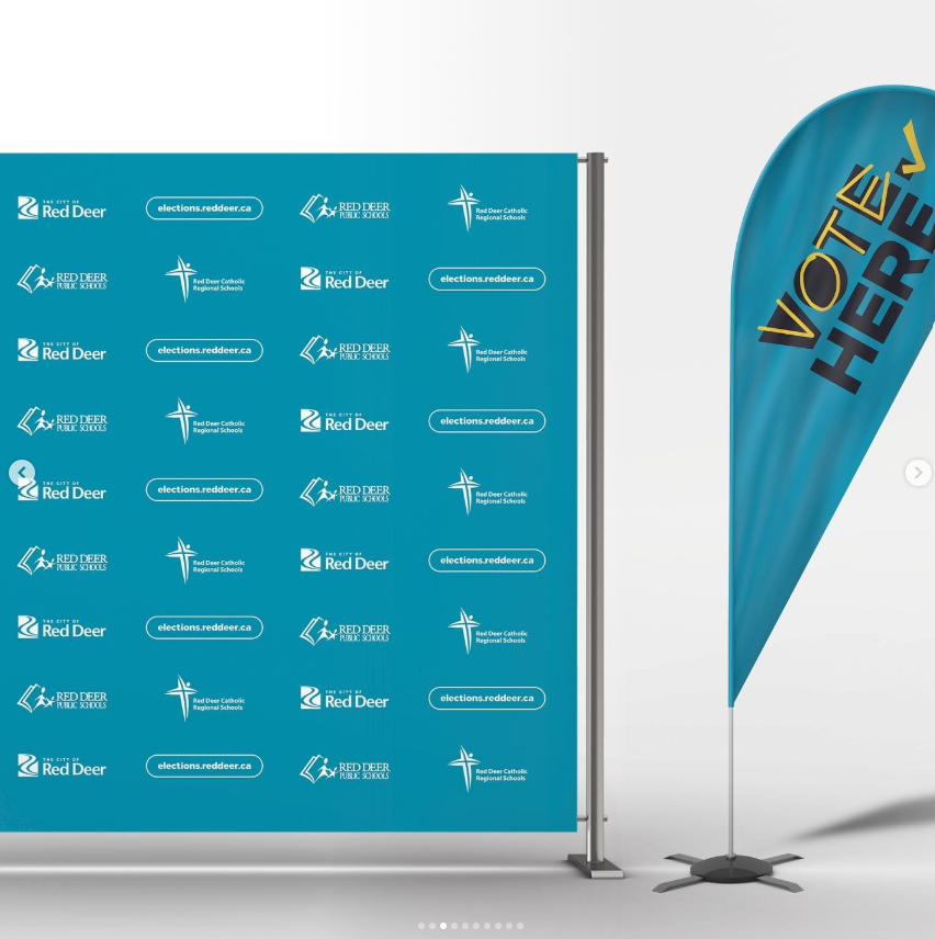A wide image showing two large election display elements for the City of Red Deer 2021 Election. On the left is a freestanding turquoise media wall backdrop mounted on a metal frame, covered in a repeating pattern of logos for the City of Red Deer, Red Deer Public Schools, Red Deer Catholic Regional Schools, and the website elections.reddeer.ca. On the right is a vertical teal feather flag on a weighted base, angled slightly, with large yellow and dark blue text reading “Vote Here.” Both elements use a consistent teal‑based colour palette designed for visibility in public or outdoor settings.
