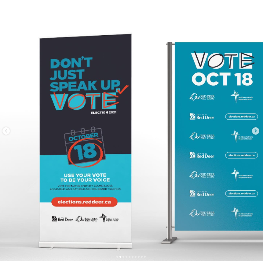 A wide image showing two freestanding election display pieces for the City of Red Deer 2021 Election. On the left is a tall vertical banner reading “Don’t just speak up, vote,” with the election year noted as 2021 and a calendar graphic highlighting October 18. Additional text reads “Use your vote to be your voice,” with references to voting for mayor, city councillors, and school board trustees, and the website elections.reddeer.ca. On the right is a turquoise media wall backdrop on a metal frame featuring large text reading “Vote Oct 18,” along with repeated logos for the City of Red Deer and local school boards. The displays share a consistent colour scheme of dark blue, teal, white, and orange accents.