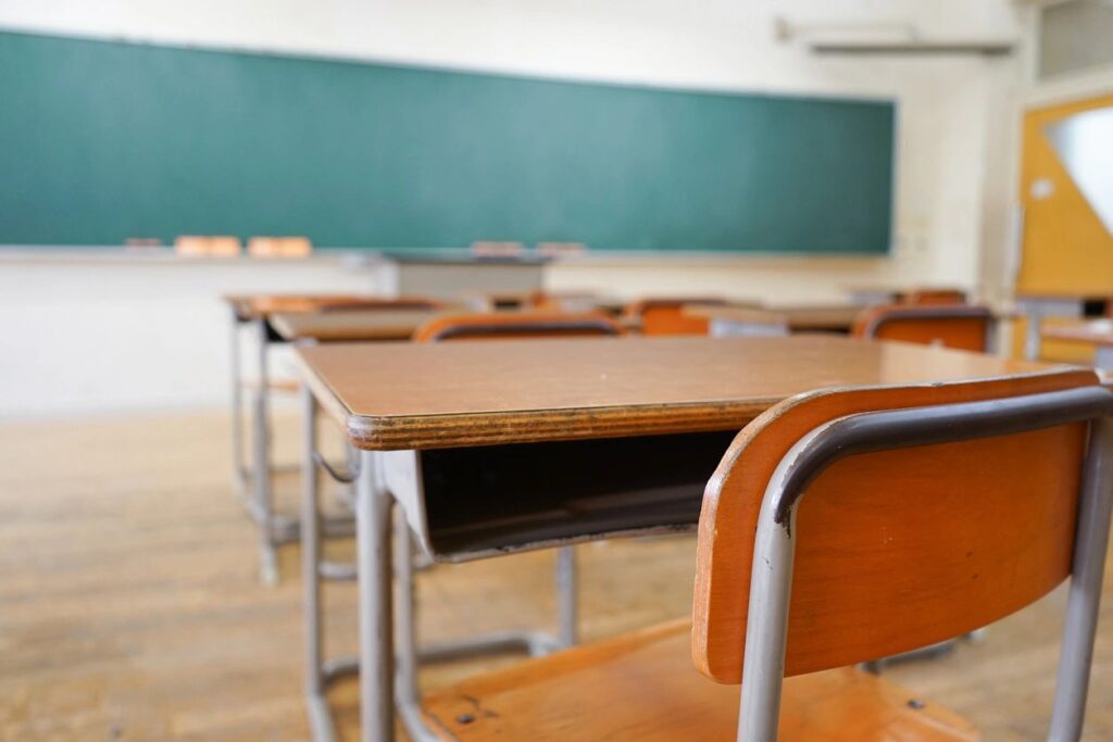 An empty classroom is shown with a empty desks in the foreground and a clean, green chalkboard in the background.